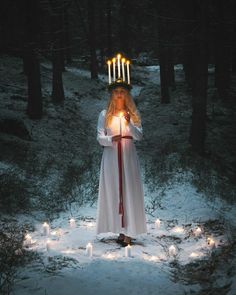 a woman dressed in white holding candles and standing in the middle of a snowy forest