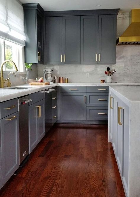 Stunning gray kitchen with gold accents boasts cherry stained oak wood floors lining gray shaker cabinets adorning brushed bras pulls and thick marble countertop fitted with a stainless steel sink and brass gooseneck faucet placed beneath a window dressed in a white roman shade. Cherry Wood Kitchen Cabinets, Cherry Wood Kitchens, Cherry Wood Cabinets, Cherry Wood Floors, Серая Кухня, Living Room Wood Floor, Wood Floor Kitchen, Oak Wood Floors, Gray Cabinets