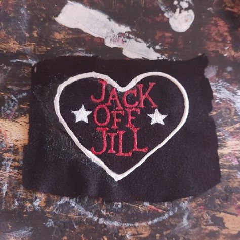 An unevenly cut, black, rectangle, fabric patch is on a paint-stained background. On the patch is a white heart, inside the heart, written in red, are the words "Jack Off Jill" in a vertical column. On either side of the word "Off" are white stars. Jack Off Jill Logo, Alternative Accessories Diy, Punk Shirts Diy, Punk Pins And Patches, Punk Patch Stencil, Punk Band Patches, Band Patches Diy, Jack Off Jill Band, Punk Diy Ideas