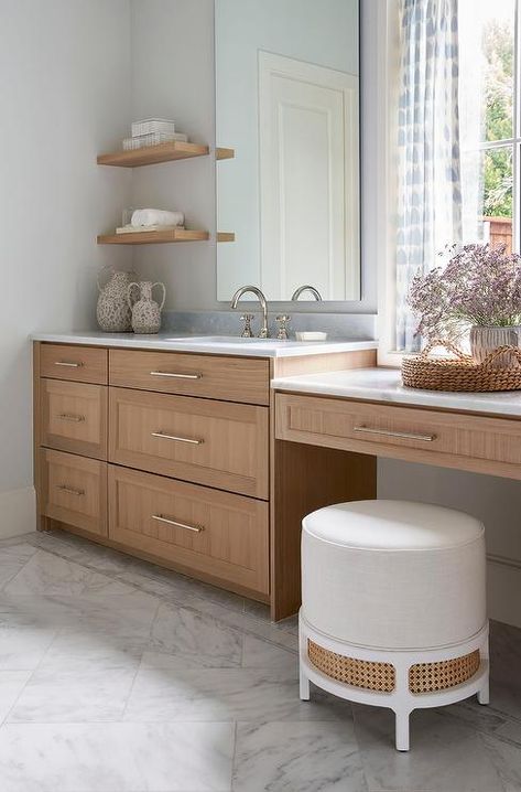 Oak floating shelves are mounted beside a frameless vanity mirror hung over a brown wooden washstand donning nickel pulls and a polished nickel gooseneck faucet fixed to a marble countertop. Beach Vanity, Flat Front Cabinets, Off White Kitchen Cabinets, Floating Kitchen Shelves, Off White Cabinets, Light Gray Cabinets, Bathroom With Makeup Vanity, Off White Kitchens, Oak Floating Shelves