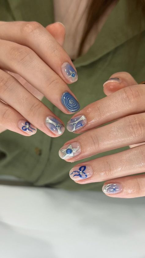 The image shows a close-up of a person's hands with an intricate blue-themed gel nail art. The designs include various patterns such as spirals, abstract shapes, stars, and bows, all in shades of blue and silver. The background is a light olive green shirt worn by the person, creating a contrasting backdrop for the nail designs. The nails have a glossy finish, highlighting the detailed and artistic work. The hands are well-manicured, and the skin appears smooth. Funky Short Acrylic Nails, Stars Nails Short, Trendy Blue Nails Short, Short Nail Designs Easy Simple, Blue Mismatched Nails, Blue And Gray Nail Ideas, Blue And Silver Short Nails, Rockstar Nail Designs, Short Art Nails