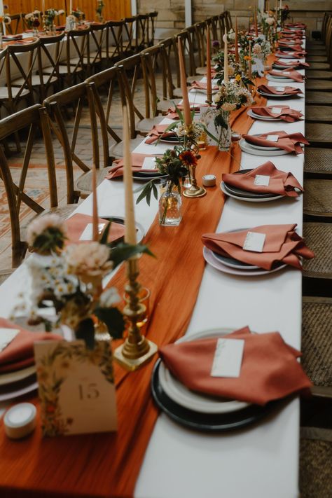 Wedding reception table decor with long burnt orange table runner and burnt orange napkins. Taper candles stand in brass candle holders & elegant Autumn flowers sit in small glass bud vases. Long trestle tables with cross back chairs. Wedding Table With Table Runner, Wedding Table Settings Burnt Orange, White And Burnt Orange Wedding Table, Burnt Orange Table Runner Wedding, Burnt Orange Table Centerpiece, Burnt Orange And Brown Wedding Decor, Rustic Wedding Decor Autumn, Diy Wedding Table Set Up, Autumn Wedding Diy Decorations