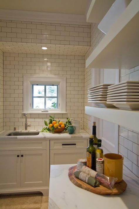 Gorgeous kitchen with white shaker cabinets accented with oil-rubbed bronze hardware, white and gray marble countertops and a ceiling height subway tiled backsplash. The kitchen sink stands below a small square shaped window across from additional cabinetry below floating white shelves. White Kitchen Renovation, Kitchen Cabinet Style, Kitchen Soffit, Small White Kitchens, White Gloss Kitchen, Small Kitchen Cabinets, White Kitchen Tiles, White Subway Tile Backsplash, White Shaker Cabinets