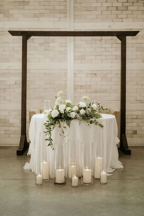 This sweetheart table is decorated with a lush floral arrangement featuring white roses, baby's breath, queen anne's lace, eucalyptus, and italian ruscus. Floral design by Imogen Floral Design. White Florals Sweetheart Table, Sweetheart Table White Flowers And Greenery, Wedding Sweetheart Table With Candles, Sweetheart Table Wedding Candles On Floor, Sweetheart Table Wedding Eucalyptus, Sweetheart Table With Eucalyptus, Head Table Wedding Decorations Candles, Minimalist Wedding Sweetheart Table, White Flower Sweetheart Table