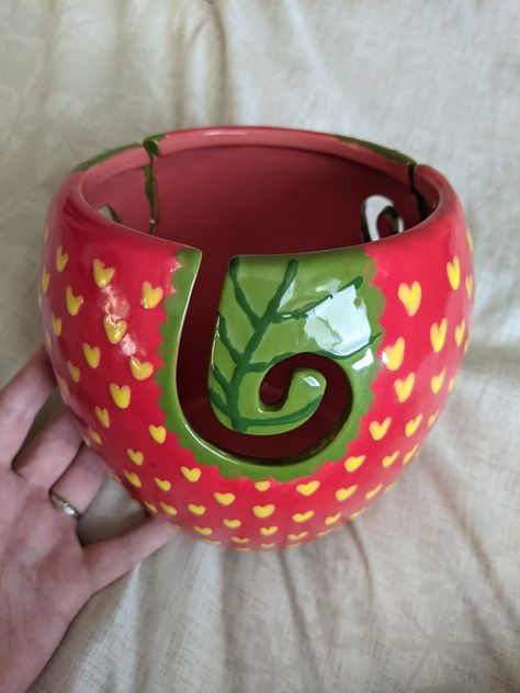 A red yarn bowl with yellow hearts painted all over it to look like a strawberry. The yarn bowl has a ceramic swirl yarn feeder that is painted green to mimic a leaf. A hand props up the bowl against a cream fabric backdrop. Ceramic Yarn Bowl Painting Ideas, Pottery Crochet Bowl, Cute Clay Yarn Bowl, Yarn Bowls Pottery Diy, Painted Yarn Bowl, Yarn Bowls Pottery Painting, Yarn Bowl Painting Ideas, Polymer Clay Yarn Bowl, Knitting Bowls Pottery