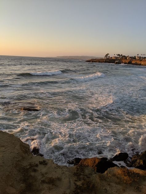 Image of the view from Sunset Cliffs in San Diego California. Image reveals a brown rocky floor in the bottom fourth, a beautiful blue and white ocean full of movement, and a few incoming new waves below. In the distance in the top right corner is a few beach houses and many palm trees that overlook the ocean. Time of day is sunset and the lighting is soft. The sky has a hint of yellow, pink, and orange on the horizon. San Diego Vision Board, Santa Monica Beach Aesthetic, Anaheim California Aesthetic, San Diego Astethic, Priscilla Core Aesthetic, San Diego Beach Aesthetic, San Diego California Aesthetic, San Diego Vibes, California Summer Aesthetic