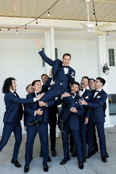 The couple wanted the wedding party to complement the environment of the venue in a timeless and upscale way that reflected the blue lake and beautiful surrounding greenery. The bridesmaids wore dusty blue Azazie dresses with soft, natural glam by LJ Artistry and Reborn Hair Company, and carried bouquets full of soft green, soft pinks, and creams. The groomsmen wore navy blue suits with black lapels and bowties from Men's Wearhouse. #lakewedding #groomsmen Wedding Groomsmen Attire Navy Blue, Cobalt Blue Groomsmen Attire, Blue Tuxes Wedding, Navy And Black Bridesmaid Dresses, Dark Blue Summer Wedding, Dark Navy Blue Groom Suit, Navy And Black Tux Wedding, Groomsmen Blue Tie, Navy Blue Bridesmaids And Groomsmen