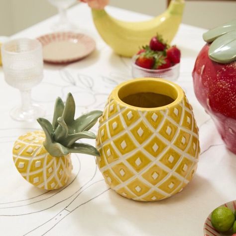 Experience the fun and fruity vibes of the Pineapple Canister! This food-safe ceramic cookie jar is adorned with vibrant hand-painted details and a yellow reactive glaze, giving it a unique and playful touch. Complete with a removable stem lid and dimensional seeds, this quirky and trendy piece is the perfect addition to your kitchen decor. Pair it with our other fruit-inspired tableware for a coordinated and summery everyday or event look. 6.5"Wx12"H Fruit Themed Kitchen, Pineapple Vase, Ceramic Pineapple, Fruit Decor, Food Shapes, Ceramic Cookie Jar, Pineapple Decor, Reactive Glaze, Candle Tray