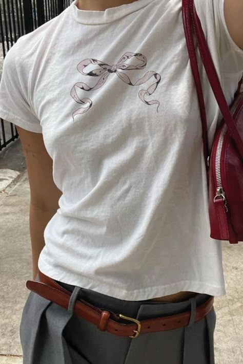 A cropped photo shows a girl from the waist to just below her neck, wearing a white tee shirt with a light pink bow illustration, paired with gray slacks, and a brown leather belt, and cherry red handbag Coquette Graphic Tee, Graphic Baby Tee Outfit, Baby Tees Outfit, Baby Tees Aesthetic, Baby Band Tees, Cute Baby Tees, Baby Tee Aesthetic, Aesthetic Ribbon, Baby Tee Outfit