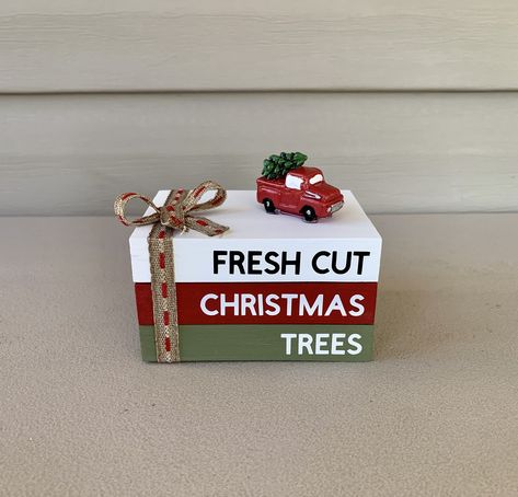 "This handmade mini wooden book stack features 3 rectangle blocks. The blocks are painted white, red, and green with permanent vinyl. The blocks display the words \" FRESH CUT CHRISTMAS TREES \" The blocks are glued together and has a red truck with Christmas tree that is also glued to the top and has a ribbon tied into a bow.  The book stack is about 3\"W x 4\"L x 2.5\" H These will ship in 3 days from ordering Thanks for shopping at  Farmhouse Charm To Love" Wooden Book Decor, Mini Book Stacks Diy, Christmas Book Stacks Diy, Christmas Book Stacks, 2 X 4 Crafts, Red Truck With Christmas Tree, Holiday Luncheon, Tiered Tray Decor Christmas, Stack Books