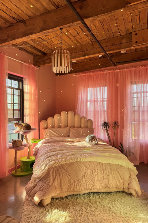 A beautifully decorated bedroom featuring a soft pink color scheme, a unique padded headboard, a cozy bedspread, sheer pink curtains, a disco ball, and a cat tree with cats perched on it. The room exudes warmth with natural light filtering through the windows and wooden ceiling beams. Pink Disco Bedroom, Angled Bed In Corner, Disco Ball Bedroom, Disco Bedroom, Decor Amazon Finds, Modern Eclectic Bedroom, Bedroom Pink, Bedroom Cozy, Velvet Upholstered Bed