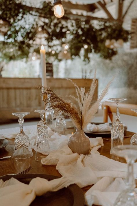 This shows a close up of a rustic barn dining tablescape. The focus is on a wooden table with white table runners on it, gold  candlesticks with LED candles, cream bud vases with dried flowers, cutlery and tableware. Dried Flower Table Decor, Dried Flower Table Arrangements, Small Table Design, Dining Table Flowers, Barn Photography, Dried Flowers Wedding, Flower Installation, Wedding Table Flowers, Wedding Arrangements