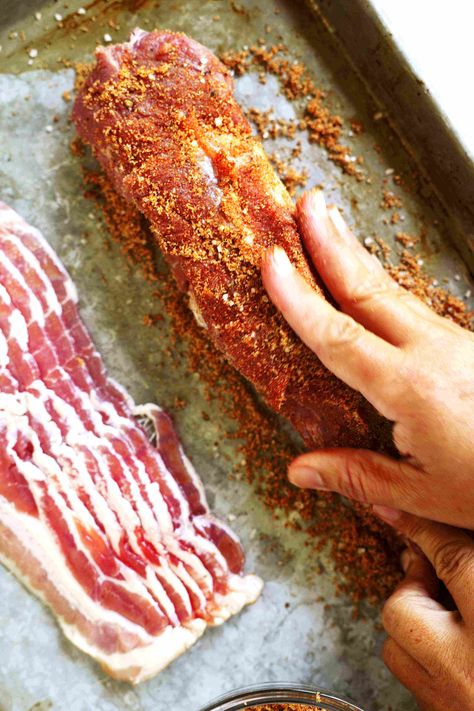 A hand rubbing the brown sugar mixture into a pork tenderloin. A stack of bacon is sitting next to it. Pork Bacon Recipe, Applewood Bacon Pork Tenderloin, Bacon Brown Sugar Pork Tenderloin, Pork Tenderloin Recipes In Oven Bbq, Pork Loin Wrapped In Bacon In Oven, Smoked Bacon Wrapped Pork Loin, Bacon Wrapped Pork Loin In Oven, Bacon Wrapped Tenderloin Pork, Pork Tenderloin Recipes In Oven Marinade