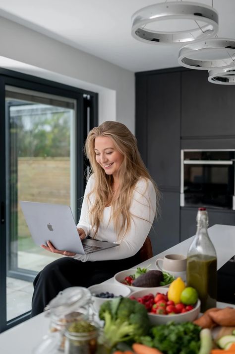 A woman with long blonde hair sits at a kitchen counter using a laptop. The counter is covered with various fruits, vegetables, and a bottle of olive oil. Large windows are seen in the background, creating an inspiring scene perfect for Liverpool brand photography or entrepreneur portraits by a UK branding photographer. Lifestyle Kitchen Photoshoot, Nutrition Coach Photoshoot, Dietitian Branding Photoshoot, Nutrition Coach Photography, Health Coaching Aesthetic, Health Wellness Photoshoot, Nutrition Photography, Nutritionist Photoshoot Health Coach, Dietician Branding Photoshoot