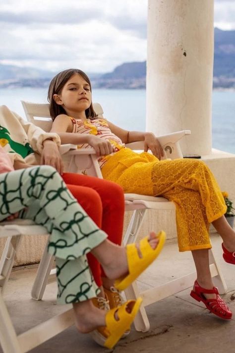 Two young girls are sitting on white outdoors chairs on a balcony overlooking a lake and hills in the back. From the right is one girl but you can't see her fully, only her legs and her left arm. Next is sitting a girl with brown straight hair and she is laying with the head back on the chair and her arms are casually placed on armrests. She is wearing bright yellow lace trousers, vibrant red shoes and a white top with red stripes and yellow seahorses. These are clothes by Mini Rodini. Lace Trousers, Children's Clothing Brand, Organic Kids Clothes, Heart Motif, Baby Clothes Patterns, Online Kids Clothes, Kids Clothes Boys, S Heart, Yellow Lace