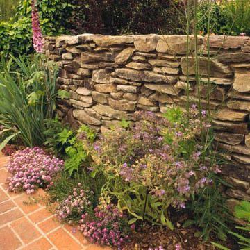 A dry-stacked stone wall looks so natural. The spaces between stones in the retaining wall allow water to drain through, relieving pressure. Another advantage of dry-stacked stone walls is that you can create planting pockets for small rock garden plants such as sedums. Garden Landscaping Design Ideas, Stone Walls Garden, Dry Stack Stone, Stacked Stone Walls, Gravel Landscaping, Backyard Garden Layout, Rock Garden Plants, Rock Garden Design, Dry Stone Wall