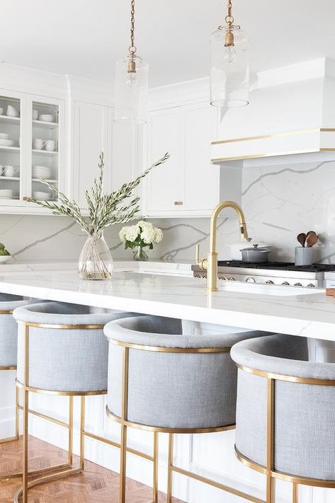 Well-appointed white and gold kitchen boasts gray linen barrel back counter stools placed on a parquet wood floor at a Calcutta marble-like island top lit by brass chain and glass pendants hung over a sink with a brushed gold faucet. White Kitchen Design, Gold Kitchen, White Modern Kitchen, Kitchen Design Decor, Kitchen Room Design, Kitchen Inspiration Design, Kitchen Stools, Kitchen Layout, Kitchen Style