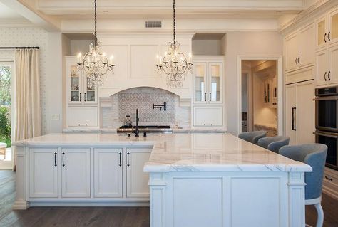 Elegant, well appointed white kitchen boasts an l-shaped white island accented with oil rubbed bronze fleur de lis pulls and a white marble countertop seating gray barrel back barstools and fitted with a sink and oil rubbed bronze faucet illuminated by two crystal chandeliers. Oil Rubbed Bronze Faucet, Kitchen Island With Sink, Sink In Island, White Island, L Shaped Kitchen, Kitchen Island With Seating, White Marble Countertops, Marble Countertop, Island With Seating