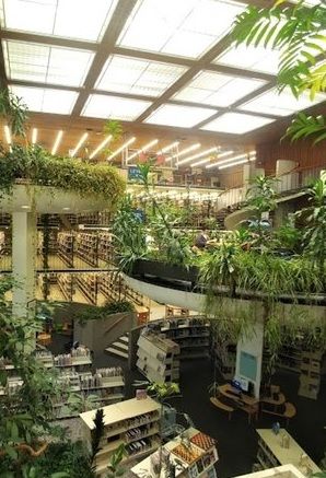 photo of the downtown Reno Library. Plant are draped over balconies and throughout the library. Large skylights are above Library In Nature, Tropical Library, Overgrown Library, Library Green, Magical Library, Green Library, Truckee River, Library Week, Library Interior