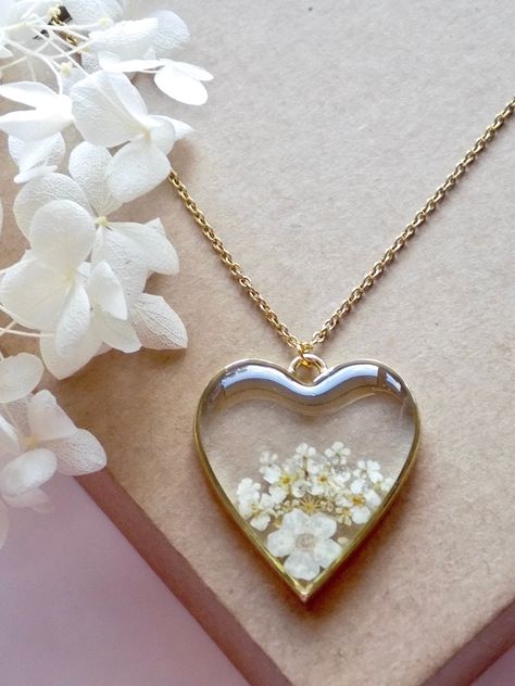 This heart shaped pendant features a variety of white garden flowers in a unique arrangement within a golden toned frame. The necklace hangs on an 18 inches long gold plated stainless steel chain. Pendant length: 35 mm Pendant width: 30mm Materials used: epoxy resin, real pressed flowers, gold plated alloy pendant frame and stainless steel chain Resin Chain Necklace, Resin Flower Pendant, Resin Necklace Ideas, Resin Jewellery Ideas, Resin Pendant Ideas, Resin Gift Ideas, Resin Art Pendant, Resin Necklace Pendant, Resin Pendant Diy