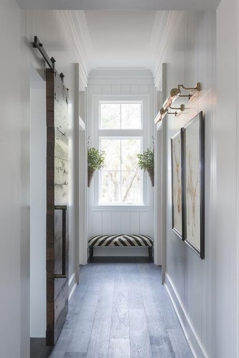 Chic hallway features vertical shiplap walls lined with side by side art illuminated by antique brass picture lights situated across from a plank barb door on rails. Narrow Hallway Lighting, Palmetto Bluff Homes, End Of Hallway, Hallway Art, Hallway Designs, Hallway Design, Upstairs Hallway, Hal Decor, Long Hallway