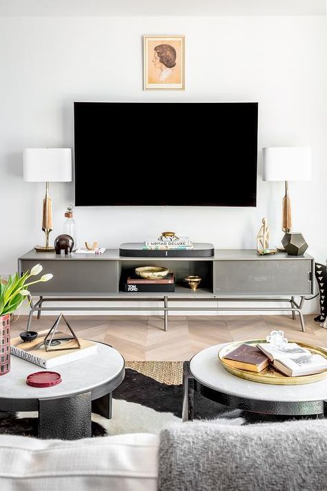 Contemporary living room boasts wood and brass lamps placed on a low gray metal tv console placed on a wood chevron floor beneath a wall mount TV fixed under a framed art piece. Metal Tv Console, Console Table Under Tv, Red Toile Curtains, Melanie Turner Interiors, Grey Wood Coffee Table, Wall Mount Tv, Mount Tv, Tv Console Modern, Under Tv