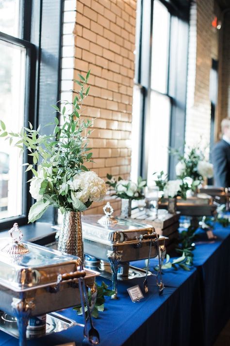 One of the last places couples think to decorate is the buffet table, but sprucing up this space will add some wow factor to your wedding. This couple used a navy linen instead of the typical black, and included some simple vases with a white hydrangea and greenery. Simple, classy, and sure to impress guests. Engagement Buffet Table, Buffet Dishes Display, Chafing Dish Display Ideas Wedding, Buffet Wedding Decor, Party Buffet Table Set Up, Rehearsal Dinner Buffet Table Decor, Buffet Centerpiece Ideas, Elegant Buffet Table Ideas Decor, Wedding Buffet Tables