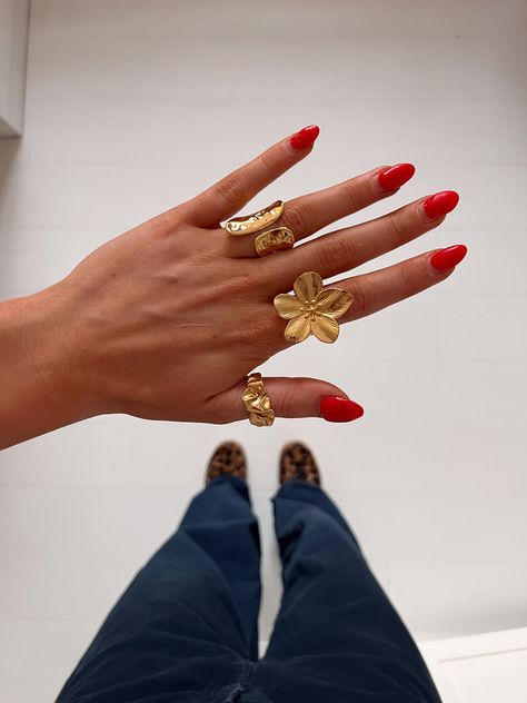 A close-up of a hand with red nail polish, wearing multiple gold rings including a flower ring, against a background of pants and leopard print shoes Ring Stacks Aesthetic, Summer Jewelry Stack, Chunky Ring Stack, Chunky Gold Jewelry, Chunky Jewelry, Dope Jewelry, Jewelry Lookbook, Stacked Jewelry, Jewelry Photography