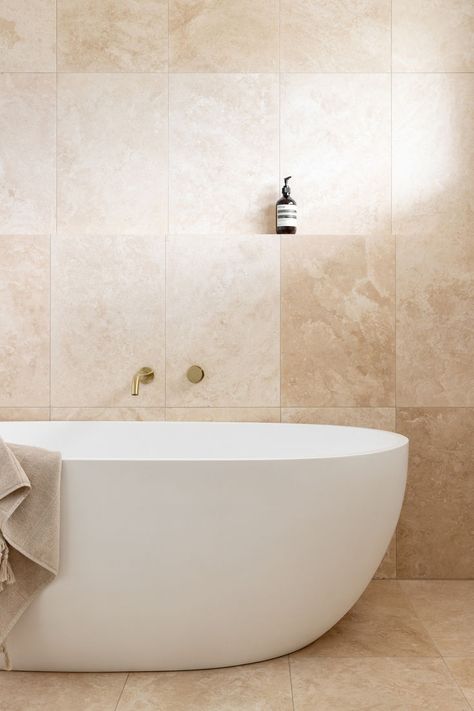 A bathroom features floor to ceiling travertine stone tiles and brushed brass tapware.