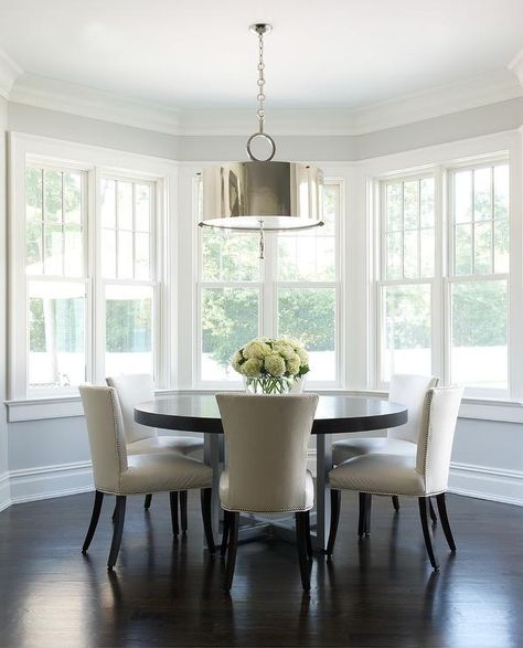 Curved dining room features a dark stained round dining table lined with ivory leather dining chairs with silver nailhead trim illuminated a polished nickel drum pendant placed in front of curved windows. Farmhouse Lighting Dining, Game Table And Chairs, Unique Dining Room, Transitional Dining Room, Round Dining Room Table, Dining Room Windows, Round Dining Room, Dining Room Table Decor, Dark Stain