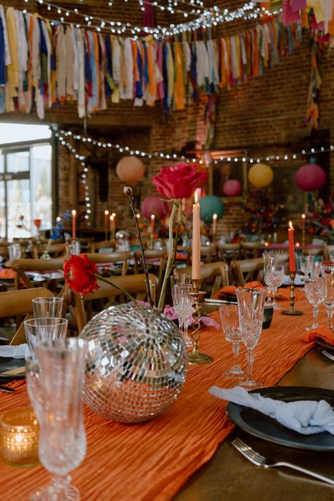 This shows a wedding dining set up. The focus is on a small silver disco ball on a dining table, the table is covered in an orange table runner. Also on the table are glassware, tableware, candlesticks. Above the tables are colourful bunting. Festival Style Wedding Receptions, Festival Wedding Table Decor, Colourful Garden Party, Colourful Wedding Ideas, Vibey Wedding, Farm Event, Love Festival, Village Hall Wedding, Wedding Setup