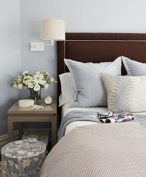 A two tier gray nightstand sits illuminated by a white swing arm sconce mounted on blue walls beside a circular blue floral print stool and a chocolate brown upholstered headboard supporting a bed dressed in a white and black duvet highlighting white shams and blue accent pillows, and a beige, blue and white reef printed lumbar pillow. Brown Headboard, Blue Headboard, Brown Decor, Black Headboard, Transitional Bedroom, Brown Bedroom, Condo Living, Black Pillows, Bedroom Black