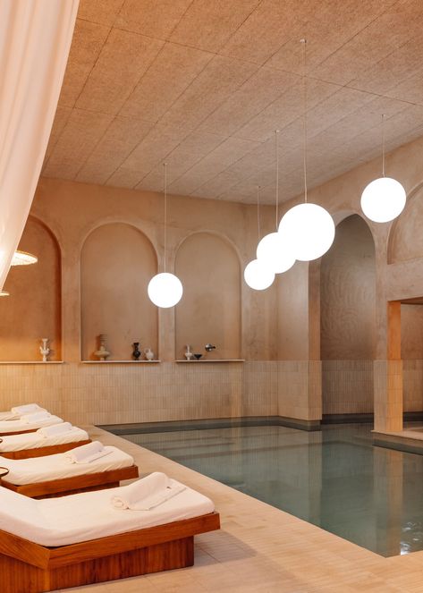 Inspired by ancient Roman thermal baths, the Spa at The Rebello is centered around serenity, calmness, and luxury, with a strong focus on wellness and relaxation. The centerpiece of the spa is an indoor pool lined with studio-designed iroko daybeds and billowing drapery. The pool is enhanced by programmed lamps that mimic the natural phases of the sun and moon, including dawn, sunset, and moonlight. Indoor Pool And Sauna, Indoor Pool Lighting, Pool House Lighting, Massage Room Lighting, Indoor Plunge Pool, Cold Plunge Spa, Interior Pool Design, Spa Pool Area, Magnesium Pool