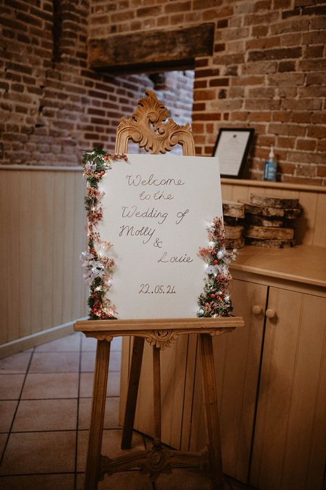 This shows a wooden ornate easel, on it is a white sign with flowers and lights around the borders. The sign reads "Welcome to the wedding of Molly x Louie 22.06.24 Photography Rugs, Flowers And Pampas, Event Signs, Barn Photography, Diwali Decorations At Home, Wood Easel, Event Signage, Wooden Easel, Event Sign