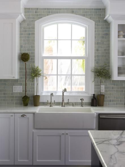 A ceramic farmhouse sink with a brushed-nickel faucet anchors this serene transitional kitchen. Countertop topiaries and a green subway-tile backsplash create an earthy backdrop for crisp white cabinetry and an arched window. Subway Tiles, Farmhouse Kitchen Backsplash, Kitchen Sink Design, Kitchen Backsplash Designs, Gorgeous Kitchens, Modern Farmhouse Kitchens, Trendy Kitchen, Kitchen Redo, Decor Minimalist