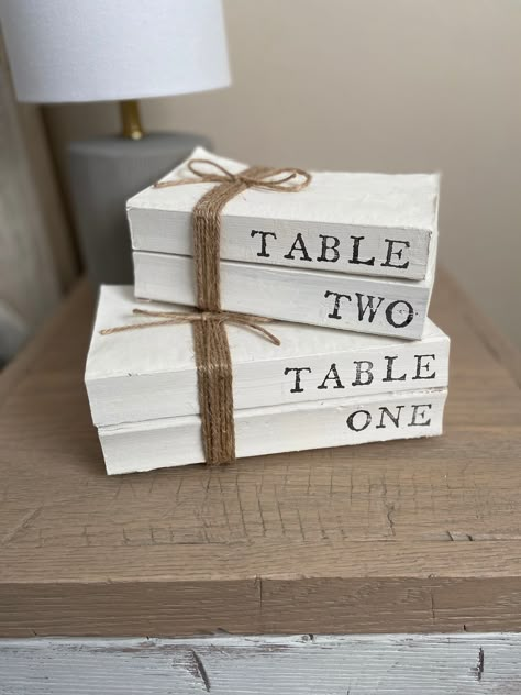 All book lovers can make their wedding days even more special with these recycle book table numbers.  These books stacks come in stacks of 2 recycled books that display the table and number.  They are also wrapped with simple twine to ensure that they are versatile to match your personal style.   These table numbers can be used beyond just your wedding day.  They also make great decor for anniversary parties, bridal showers, or baby showers.  These stacks are made from recycled books looking for a new chance at life.  Variations on the spine or pages may be present since these books have been loved by many literature lovers.  They are painted and hand stamped to achieve a rustic appearance.  Standard book stacks are painted white with black lettering; however, colors can be personalized at Wedding Table Book Centerpieces, Book Bridal Shower Theme Centerpieces, Wedding Ideas Book Theme, Book Wedding Aesthetic, Book Theme Wedding Decorations, Wedding Decor Books, Bridal Shower For Book Lover, Wedding Book Theme, Bookish Wedding Decor