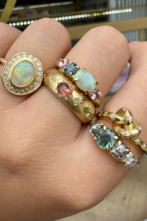 Close-up of a hand wearing five yellow-gold rings, each with a different style and type of gemstone. Two have a row of four claw-set stones, one is a 'pretzel' knot ring studded with white diamonds, one is a wide band with inset gemstones and  the last has a large central oval opal with a white diamond halo. Family Heirloom Jewelry, Five Gold Rings, Gold And Light Blue, Famous Jewelry, Colorful Ring, Jewelry Big, Chic Earrings, Dope Jewelry, Stylish Bracelet