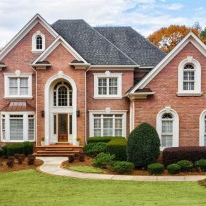 Red brick house with dark gray roof shingles. Medium brown wood stained front door with white transoms and matching stone white trim. Black metal accent roofing. Red Brick House Shingle Colors, Grey Roof Red Brick House, Red Brick House Exterior Roof Color, Red Brick House With White Trim, Houses With Gray Roofs, Shingles For Red Brick House, Red Brick House With Black Roof, Red Brick House White Trim, Black Roof Red Brick House