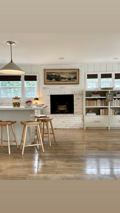 Kitchen renovation, fireplace in kitchen, limewashed fireplace, 1950 beach house, kitchen island in front of fireplace, library in kitchen, builtin shelves in kitchen, wooden woven shades, shiplap walls, swiss coffee paint on walls, laminate wood floors, winds breath paint benajmin more, pendants above kitchen island, wooden kitchen stools, art above fireplace, modern traditional home, art in front of shelves, thrifted home, tiny windows, painted fireplace, books turned around, shelf styling Kitchen Next To Fireplace, Kitchen Brick Fireplace, Fireplace Next To Kitchen, Fireplaces In Kitchen, Kitchen With A Fireplace, Kitchen Design With Fireplace, Fireplace In Kitchen Ideas, Exposed Brick Fireplace Kitchen, Old Fireplace In Kitchen