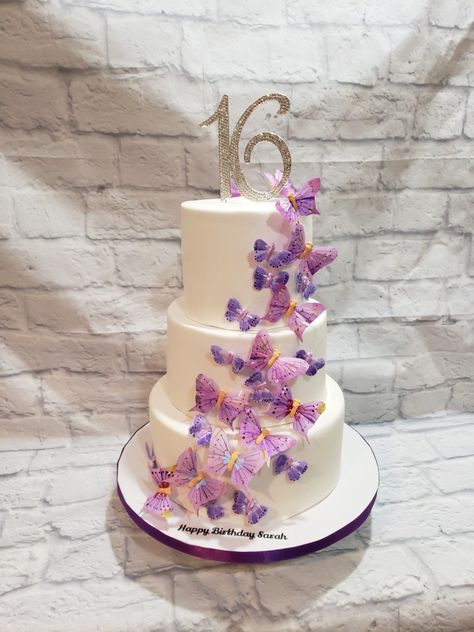 Three tiered Sweet 16 cake in all white fondant with  cascading  butterflies in different shades of purple with 16 rhinestone topper Sweet 16 Cakes With Butterflies, Sweet 15 Butterfly Cake, Sweet 16 Cake Butterfly, Butterfly Themed Birthday Party Sweet 16, Purple Sweet 16 Dessert Table, 3 Tier Cake Birthday Sweet 16, Purple And White Butterfly Cake, Butterfly Party Ideas Sweet 16, Purple And White Sweet 16 Cake