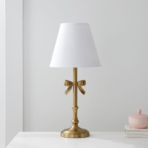 Tie their room together with this charming table lamp. It's expertly hand finished to emulate brushed gold and it comes topped with a white linen shade to cast soothing, filtered light around the room. DETAILS THAT MATTER Expertly crafted with aluminum and steel. Lamp base features a ribbon and is hand finished in a water-based Brushed Gold. On/off switch located on in-line; white cord. Shade is included and exterior is made from linen. Interior is lined with styrine. All our shades are designed White And Gold Table Lamp, Gold Bow Lamp, Cute Gold Lamp, Vintage Modern Room Decor, Silver And Gold Room Decor, Pink Lamp Shades, Trinkets For Room, White And Gold Lamps, Cute Small Lamps