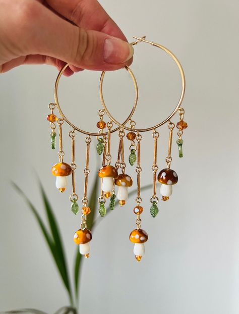 Super Cute Dangle Mushroom Hoop Earrings. These earrings feature gold hoops adorned with dangling mushrooms, accented by orange beads and small green leaves for a whimsical and nature-inspired look. Beaded Crystal Earrings, Hippy Earrings, Earrings Mushroom, Whimsical Earrings, Cottagecore Earrings, Mushroom Jewelry, Mushroom Earrings, Whimsical Jewelry, Funky Earrings