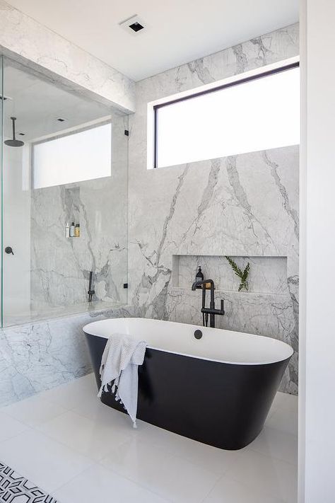 Elegant bathroom boasts a modern black and white oval bathtub with a matte black floor mount tub filler positioned on a white tiled floor beneath a marble tiled niche framed by a bookmatched marble slab wall finished with a picture window. Window Above Bathtub, Tub Niche, Tiled Bathtub, Tiled Niche, Shampoo Niche, Tub Bathroom Ideas, Transitional Bathroom Design, Concrete Bathtub, Country Style Bathrooms