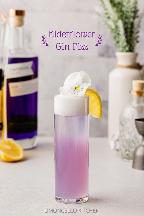 Side view of purple-colored Elderflower Gin Fizz cocktail with a lemon and edible flower as garnish and ingredients in the background. Buzz Button Cocktail Recipe, Beginner Cocktails, Upscale Cocktails, Gin Fizz Recipe, Empress 1908 Gin, Elderflower Cocktail, Mixology Recipes, Gin Fizz Cocktail, Gin Cocktail Recipes