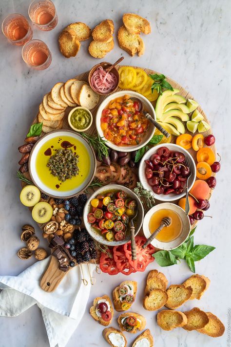 A wooden board with bruschetta toppings (pickled cherries, marinated tomatoes, apricot salsa, olive oil dip), fruit, nuts, avocado, pesto, tomatoes and chocolate. Around the board are some toasted slices of baguette. Everything is on a marble surface. Apricot Salsa, Olive Oil With Herbs, Marinated Cherry Tomatoes, Bruschetta Board, Dipping Oil Recipe, Olive Oil Dip, Pickled Cherries, Marinated Tomatoes, Charcuterie Recipes