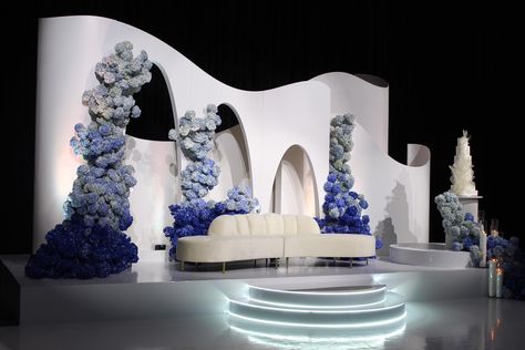 An ethereal white stage adorned with blue and white hydrangeas, a modern curved backdrop, and a white velvet love seat, creating a luxurious ambiance for a wedding reception. Modern Wedding Stage Design, Reception Stage Decoration Backdrops, Modern Wedding Backdrop, Modern Arches, Blue And White Hydrangea, Prewedding Shoot, Wedding Stage Backdrop, Reception Backdrop, Wedding Stage Design