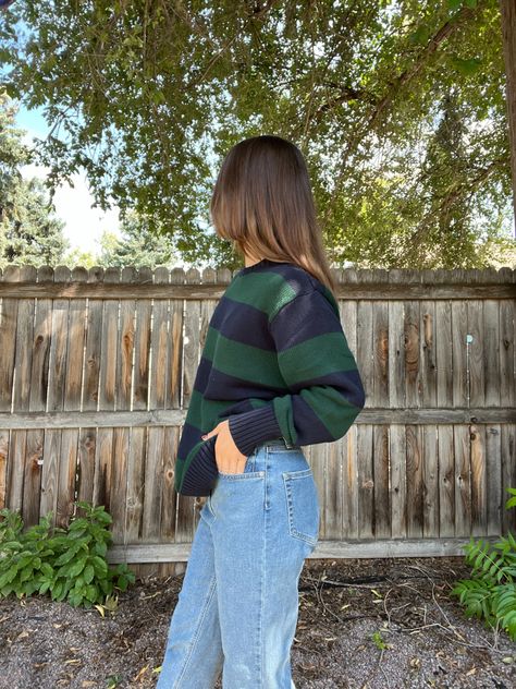 Woman standing in front of a weathered brown fence, wearing light wash baggy jeans and a sweater with large green and navy blue stripes. She is not looking at the camera and is facing sideways (left). There are green trees and plants in the background and the sun is peeking through the tree leaves in the top right corner Jean Sweater Outfit, Baggy Jumper Outfit, Jumper And Jeans Outfit, Sweater Jeans Outfit, Jeans And Sweater Outfit, Baggy Sweater Outfits, Levi Jeans Outfit, Jeans Fall Outfit, Jumper Outfits