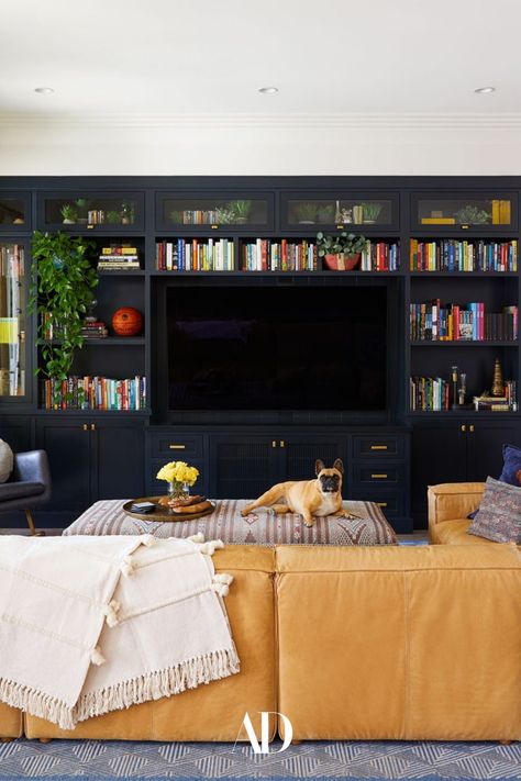 Inspired by an awakening of wanting a home that represented who they truly are, color was quickly allowed to reign supreme in the home of actors Daveed Diggs and Emmy Raver-Lampman. In the living room, the couple’s French bulldog, chills on the family room’s custom-made ottoman. #livingroom #livingroominspo #livingroomideas #familyroom #sofa #couch #leather #ottoman #entertainmentcenter #puppy #rug #curtains #sectional #books #floorlamp Build In Sofa Ideas, Tv Room With L Shaped Sofa, Architectural Digest Family Room, Leather Couch With Fabric Ottoman, Media Area Living Room, Daveed Diggs House, Unique Family Room Ideas, Family Room Leather Sectional, Home Office With Sectional