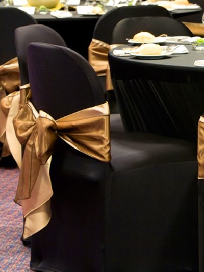 Black spandex chair cover with simple gold sash! Chair Covers Wedding Reception, Black Chair Covers, Golden Chair, Wedding Chair Covers, Black And Gold Party Decorations, Chair Covers Party, Batman Wedding, Golden Table, Gold Sash