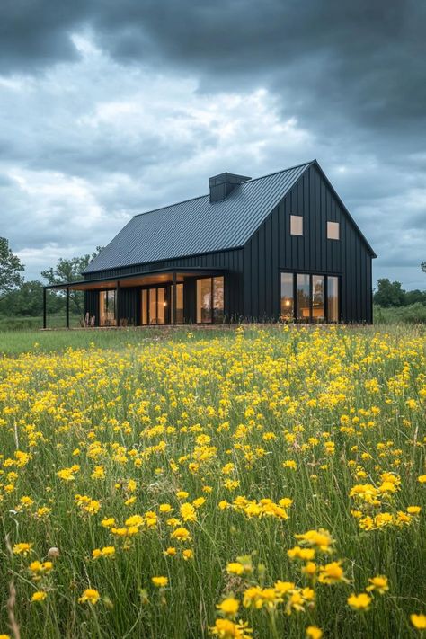 Modern barndominium charcoal black facade in stunning farmhouse meadow with yellow flowers. Check out the coolest dark barndominium houses that combine rustic charm with modern sophistication, enhancing a living space with bold hues and sleek designs. Contemporary Barndominium Ideas, Barndominium With Screened In Porch, Barndominium Landscaping, Steel Homes Metal Houses, Dark Barndominium Exterior, Dark Barndominium, Mountain Barndominium, Black Barndominium Exterior, Contemporary Barndominium